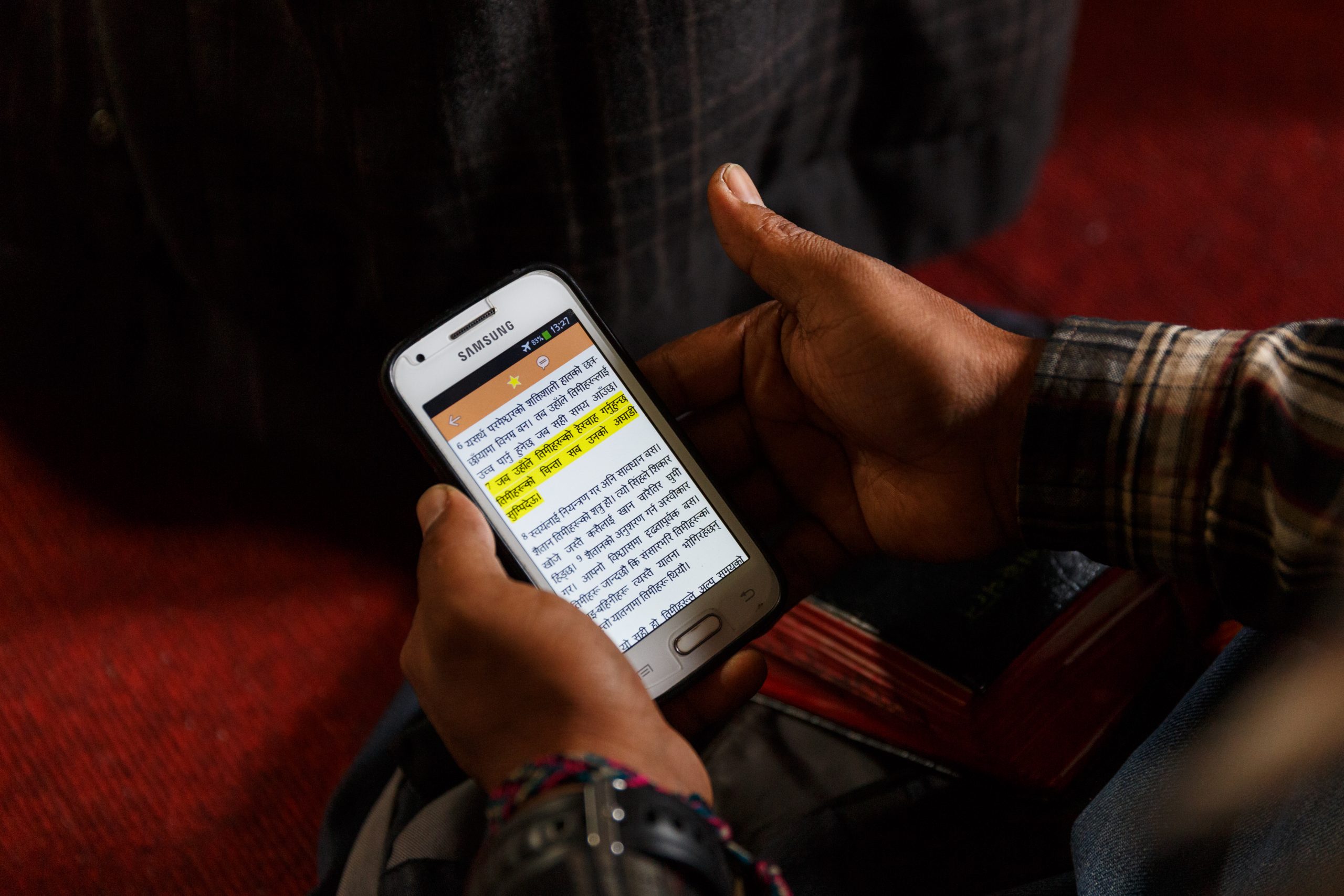 Hands holding Samsung phone with devanagari script displayed and a highlighted verse.