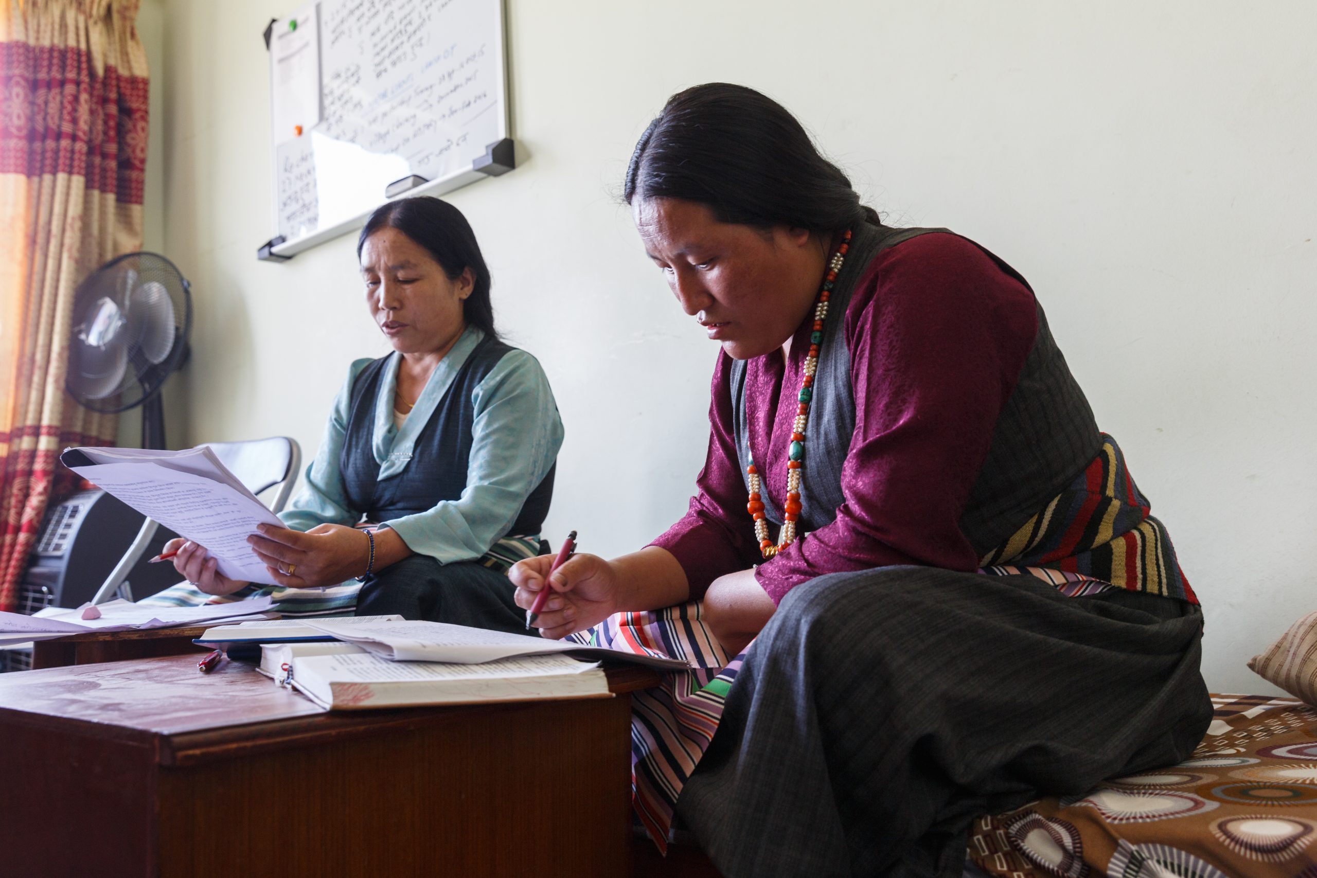 Two women hold papers and pens, sitting at a short wooden table with books open.