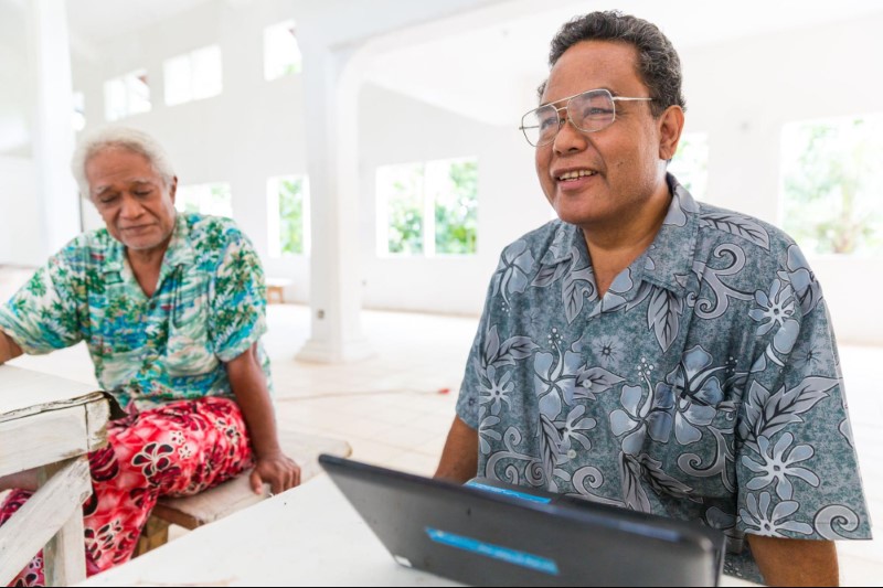 A man wearing glasses and a blue patterned shirt sits in front of an open laptop.